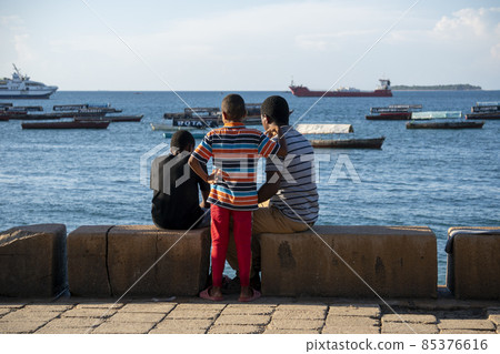 Black African Guys are sit on the Embarkment and Look in the Sea with boats in Stone Town, Zanzibar, Tanzania 85376616