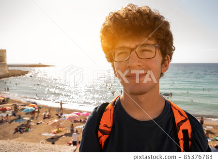 Gallipoli, Puglia, Italy. Portrait of a cute Caucasian boy with glasses and curly hair, behind the sandy beach of the old town is full of people bathing and sunbathing. 85376783