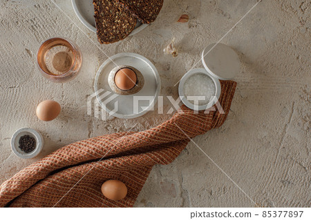 Breakfast setting with boiled egg in stone egg cup, modern concrete table accessories, whole grain rye bread with seeds, glass of water, salt flakes and pepper in concrete bowls on rough textured clay Breakfast setting with boiled egg in stone egg cup, modern concrete table accessories, whole grain rye bread with seeds, glass of water, salt flakes and pepper in concrete bowls on rough textured clay 85377897