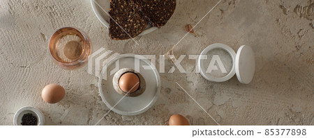 Breakfast setting with boiled egg in stone egg cup, modern concrete table accessories, whole grain rye bread with seeds, glass of water, salt flakes and pepper in concrete bowls on rough textured clay Breakfast setting with boiled egg in stone egg cup, modern concrete table accessories, whole grain rye bread with seeds, glass of water, salt flakes and pepper in concrete bowls on rough textured clay 85377898