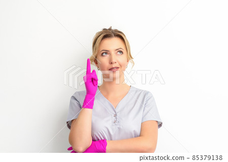 Portrait of young caucasian doctor woman wearing rubber gloves in medical uniform with one finger up, looking up against a white background, copy space. Portrait of young caucasian doctor woman wearing rubber gloves in medical uniform with one finger up, looking up against a white background, copy space. 85379138