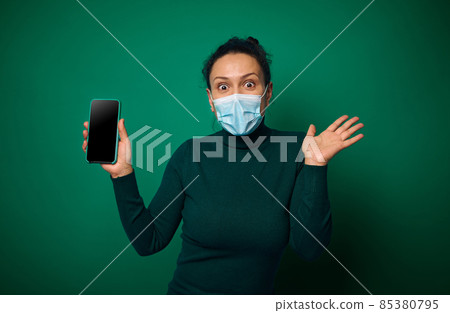 Cheerful happy millennial woman in protective face cover looks at camera and shows her smartphone with empty blank screen for copy space, isolated over green background Cheerful happy millennial woman in protective face cover looks at camera and shows her smartphone with empty blank screen for copy space, isolated over green background 85380795