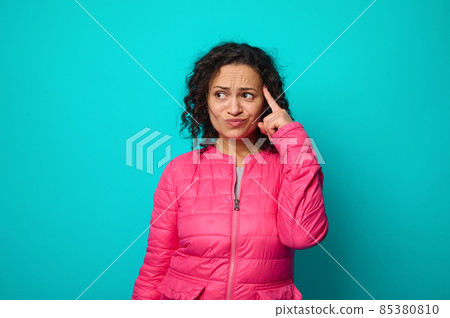 Pensive puzzled thoughtful curly haired woman in bright pink jacket holding her finger on her temple and looking thoughtfully aside to a copy space for advertising text, isolated on blue background Pensive puzzled thoughtful curly haired woman in bright pink jacket holding her finger on her temple and looking thoughtfully aside to a copy space for advertising text, isolated on blue background 85380810