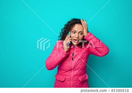 Astonished shocked emotional curly haired beautiful woman in bright pink jacket talks on mobile phone, and gestures putting her hand on her head , isolated on blue colored background with copy space Astonished shocked emotional curly haired beautiful woman in bright pink jacket talks on mobile phone, and gestures putting her hand on her head , isolated on blue colored background with copy space 85380818
