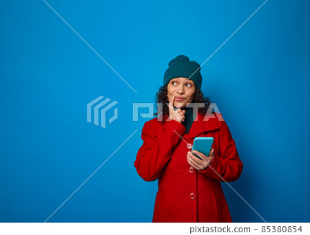 Conceptual portrait of a puzzled pensive pretty woman wearing bright red coat and warm green woolen hat holding smartphone and looking thoughtfully on a copy ad space , isolated on blue background 85380854