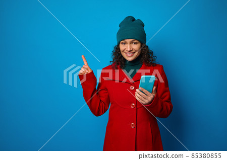 Conceptual portrait of a cheerful pretty woman wearing bright red coat and warm green woolen hat holding mobile phone and pointing with her finger on a copy space for ads, isolated on blue background Conceptual portrait of a cheerful pretty woman wearing bright red coat and warm green woolen hat holding mobile phone and pointing with her finger on a copy space for ads, isolated on blue background 85380855