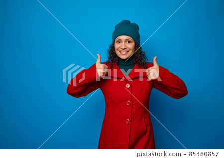 Charming wonderful attractive pretty woman in bright red coat and green hat smiling with cheerful toothy smile, looking at camera and showing thumbs up, posing against blue background with copy space Charming wonderful attractive pretty woman in bright red coat and green hat smiling with cheerful toothy smile, looking at camera and showing thumbs up, posing against blue background with copy space 85380857