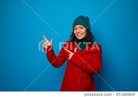Charming smiling curly dark-haired woman in bright red coat and woolen green hat poses against blue background, looks at camera and points with fingers on imaginary copy space on her hands palms up 85380869