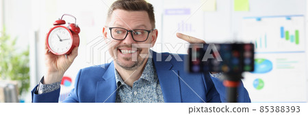 Young male blogger shoots video witha red alarm clock on camera Young male blogger shoots video witha red alarm clock on camera 85388393