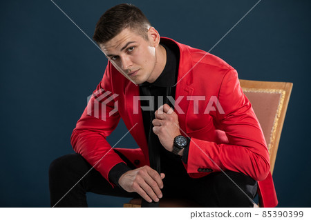 A young elegant man in a red blazer and black shirt, is improving his tie while sitting in a chair. 85390399