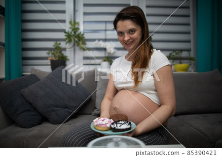 A pregnant woman sits on the sofa at home and plans to eat some donuts. A woman in advanced pregnancy. A pregnant woman sits on the sofa at home and plans to eat some donuts. A woman in advanced pregnancy. 85390421