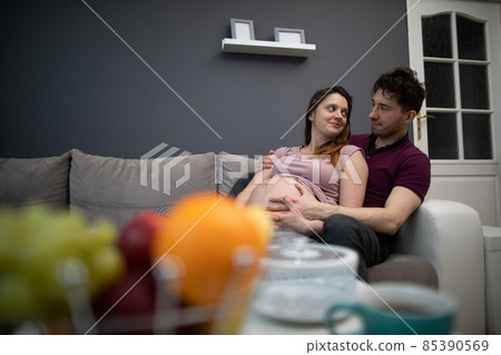 A married couple looks at each other with great joy while sitting on the sofa in the living room. The woman is pregnant. A married couple looks at each other with great joy while sitting on the sofa in the living room. The woman is pregnant. 85390569