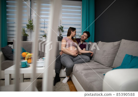 A husband hugs his pregnant wife. A married couple is sitting on a sofa in the living room at home. The pregnant woman. A husband hugs his pregnant wife. A married couple is sitting on a sofa in the living room at home. The pregnant woman. 85390588