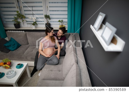 A husband hugs his pregnant wife. A married couple is sitting on a sofa in the living room at home. The pregnant woman. 85390589