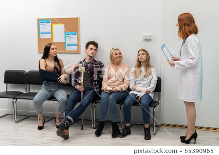 The doctor standing in front of her office reads out the next person and invites them in for a check-up. People of different age are sitting on chairs The doctor standing in front of her office reads out the next person and invites them in for a check-up. People of different age are sitting on chairs 85390965