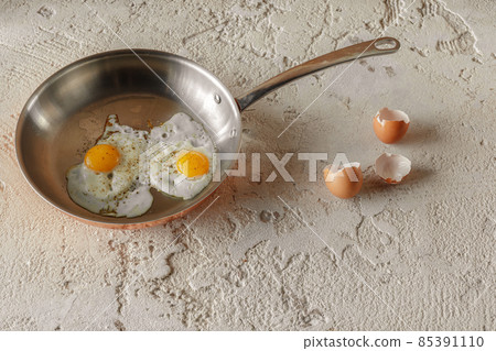 Two fried eggs in copper pan and egg shells on rough textured clay background. Healthy breakfast concept. Two fried eggs in copper pan and egg shells on rough textured clay background. Healthy breakfast concept. 85391110