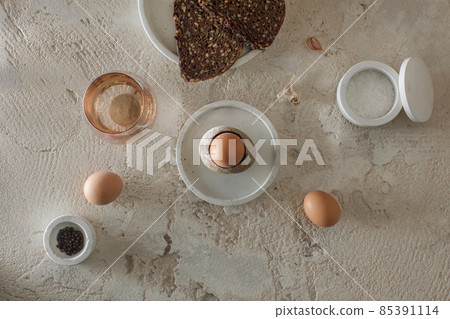 Breakfast setting with boiled egg in stone egg cup, modern concrete table accessories, whole grain rye bread with seeds, glass of water, salt flakes and pepper in concrete bowls on rough textured clay Breakfast setting with boiled egg in stone egg cup, modern concrete table accessories, whole grain rye bread with seeds, glass of water, salt flakes and pepper in concrete bowls on rough textured clay 85391114