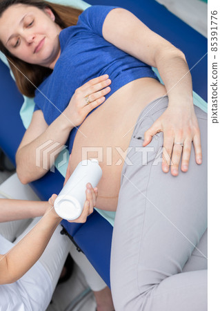 A nurse squeezes gel onto a pregnant patient's abdomen and prepares for an ultrasound. Gynecology office. 85391776