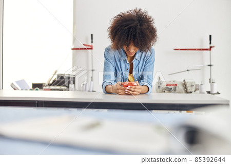 Young African-American woman surfs internet with phone at cutting table in sewing workshop Young African-American woman surfs internet with phone at cutting table in sewing workshop 85392644