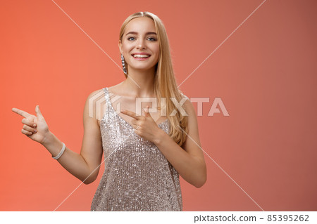 Proud happy good-looking stylish blond attractive caucasian woman in silver luxurious glamour dress pointing left smiling broadly showing friends new apartment, standing red background Proud happy good-looking stylish blond attractive caucasian woman in silver luxurious glamour dress pointing left smiling broadly showing friends new apartment, standing red background 85395262