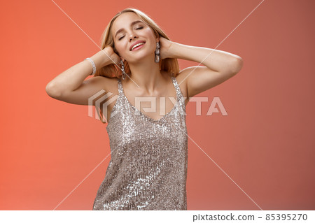 Carefree relaxed feminine lovely blond woman in silver dress enjoying slow music dance-floor run hands through hair feel free relieved joyful close eyes raise head enjoying chilling vibe 85395270