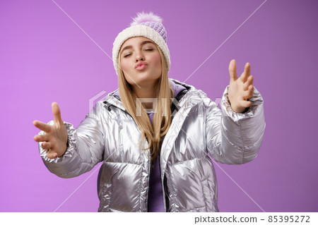 Romantic charming caucasian blond woman in cute hat silver stylish jacket pulling arms forward wanna embrace tight cuddle boyfriend folding lips close eyes give kiss, standing purple background 85395272