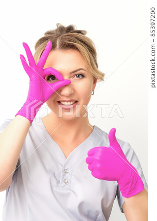Smiling doctor oculist caucasian woman wearing pink rubber gloves in uniform showing ok sign covering the eye and thumb up gesture against a white background. 85397200