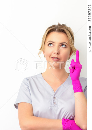 Portrait of young caucasian doctor woman wearing rubber gloves in medical uniform with one finger up, looking up against a white background, copy space. Portrait of young caucasian doctor woman wearing rubber gloves in medical uniform with one finger up, looking up against a white background, copy space. 85397201