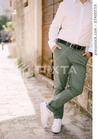 Man in green trousers and a white shirt is leaning against the wall of a stone house. Close-up Man in green trousers and a white shirt is leaning against the wall of a stone house. Close-up 85400559