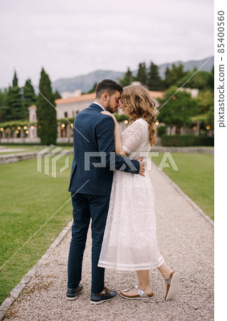 Groom hugs bride in a white dress while standing on a path among the garden on the background of an old villa and green trees Groom hugs bride in a white dress while standing on a path among the garden on the background of an old villa and green trees 85400560