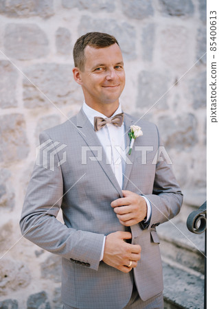 Groom holds the lapels of his jacket with his hands and stands on the stone stairs 85400613