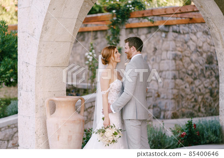 Bride and groom with a bouquet stand under a beautiful stone arch 85400646