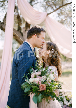 Groom kisses bride on the forehead with a bouquet of flowers under a tree. Portrait 85400727