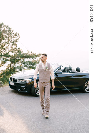 Groom walks along the road from the convertible 85401041