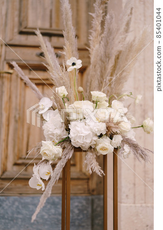 Bouquet with a cortaderia and white flowers stands on a gold pedestal in front of a wooden door Bouquet with a cortaderia and white flowers stands on a gold pedestal in front of a wooden door 85401044