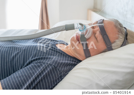 Middle age man wearing CPAP mask and headgear to help with his sleep apnea while sleeping in his bed in his bedroom 85401914