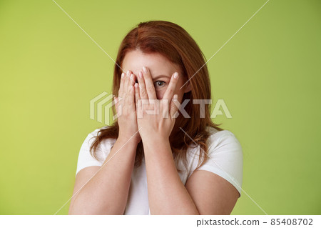 Not peeking. Playful charismatic middle-aged ginger redhead woman close face palms look through fingers one eye stare camera intrigued waiting surprise gift stand green background 85408702