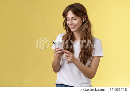 Happy caring tender feminine girlfriend curly long hair hold smartphone picking song listen way home smiling broadly look mobile phone scroll music platform wear wireless earbud yellow background Happy caring tender feminine girlfriend curly long hair hold smartphone picking song listen way home smiling broadly look mobile phone scroll music platform wear wireless earbud yellow background 85409928