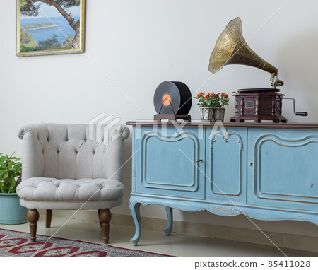 Retro off white armchair, vintage wooden light blue sideboard, old phonograph (gramophone) and vinyl records on background of beige wall, tiled porcelain floor, and red carpet Retro off white armchair, vintage wooden light blue sideboard, old phonograph (gramophone) and vinyl records on background of beige wall, tiled porcelain floor, and red carpet 85411028