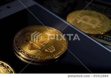 Physical Gold Bitcoin Coin against dollar bills and smartphone on a purple background. 85412664