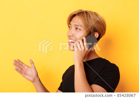 Close-up of attractive korean guy smiling and talking on mobile phone happy, turning left, standing yellow background 85428067
