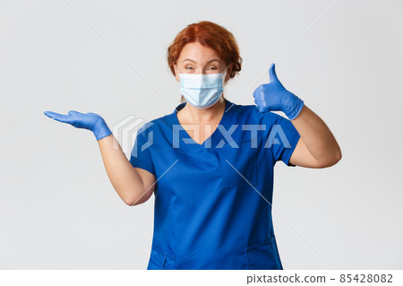 Medical workers, covid-19 pandemic, coronavirus concept. Happy smiling female doctor, vet or physician in face mask and gloves, holding something on palm and show thumbs-up in approval, recommend 85428082