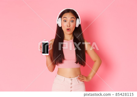Technology, communication and online lifestyle concept. Impressed asian girl found awesome podcast, showing smartphone screen as wearing headphones, listening music, pink background Technology, communication and online lifestyle concept. Impressed asian girl found awesome podcast, showing smartphone screen as wearing headphones, listening music, pink background 85428115