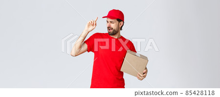 Order delivery, online shopping and package shipping concept. Carefree handsome bearded courier in red uniform, cap and t-shirt, holding box package, singing song, grey background 85428118