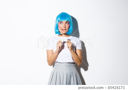 Image of thoughtful cute asian girl in blue wig making choice, looking upper left corner and showing credit card 85428171