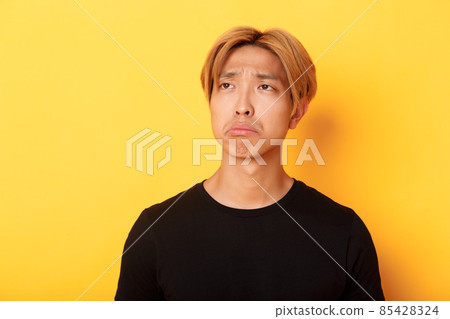 Close-up of sad and disappointed handsome asian guy sulking upset, looking upper left corner with regret or jealousy, standing yellow background Close-up of sad and disappointed handsome asian guy sulking upset, looking upper left corner with regret or jealousy, standing yellow background 85428324