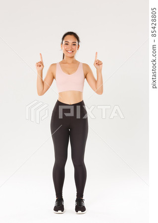 Sport, gym and healthy body concept. Full length of cheerful smiling and cute fitness girl, pointing fingers up, showing workout equipment advertisement, buy membership for training Sport, gym and healthy body concept. Full length of cheerful smiling and cute fitness girl, pointing fingers up, showing workout equipment advertisement, buy membership for training 85428805