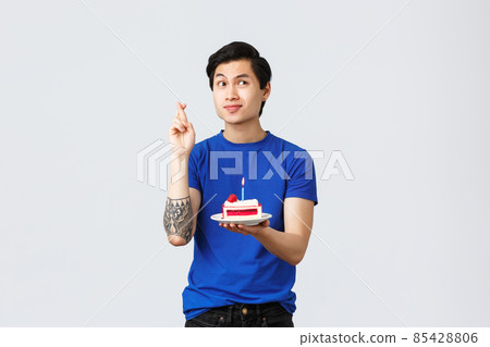 Self-quarantine, home lifestyle and celebration concept. Thoughtful dreamy asian guy, cute man celebrating birthday, cross fingers making wish and holding b-day cake with lit candle Self-quarantine, home lifestyle and celebration concept. Thoughtful dreamy asian guy, cute man celebrating birthday, cross fingers making wish and holding b-day cake with lit candle 85428806