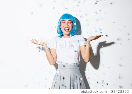 Portrait of happy asian girl in blue wig celebrating halloween, throwing confetti in the air, standing over white background 85428833