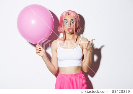 Image of excited attractive girl blowing bubble gum, showing thumbs-up in approval, standing with cute pink balloon while wearing party outfit and wig Image of excited attractive girl blowing bubble gum, showing thumbs-up in approval, standing with cute pink balloon while wearing party outfit and wig 85428859